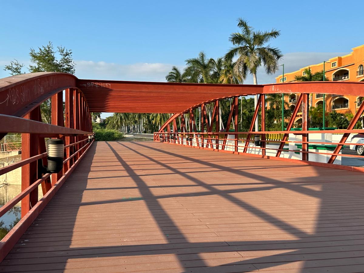 A red steel pedestrian bridge with arched trestle supports crossing a freshwater stream in Huatulco, Mexico, framed by palm trees